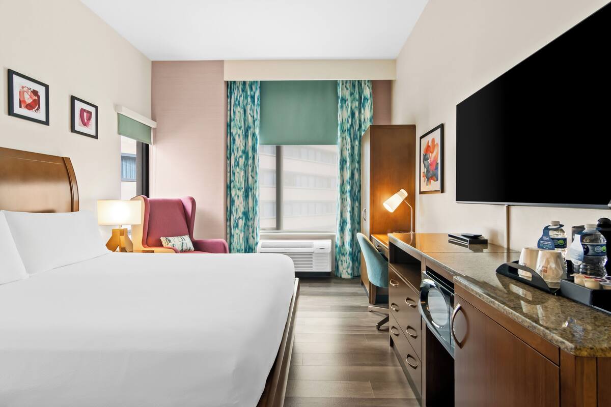 A hotel room with a bed, desk, chair, television, and a window with curtains.