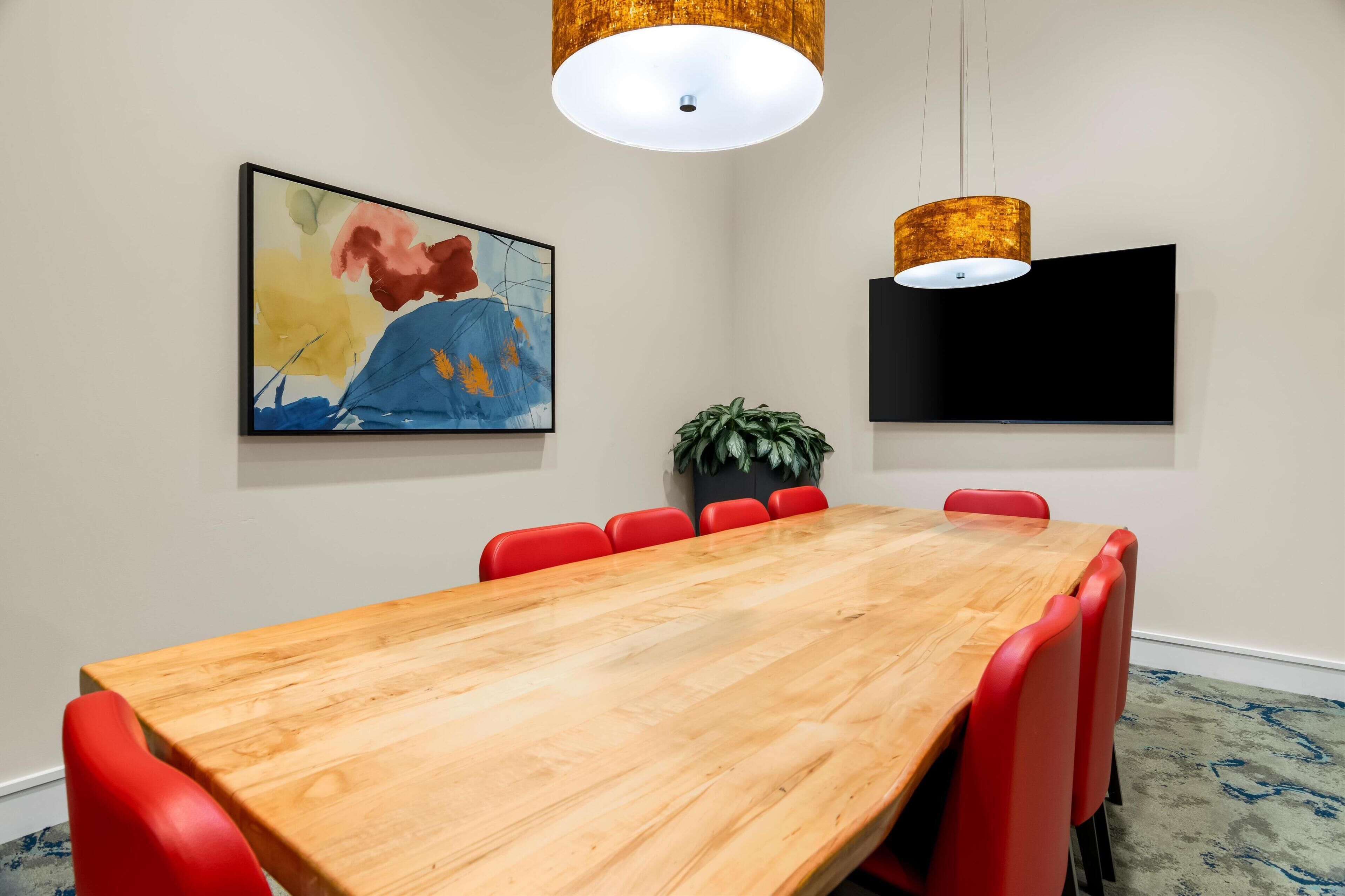 Uma sala de conferências com uma mesa de madeira, cadeiras vermelhas, um grande quadro abstrato, uma TV de tela plana e um lustre.