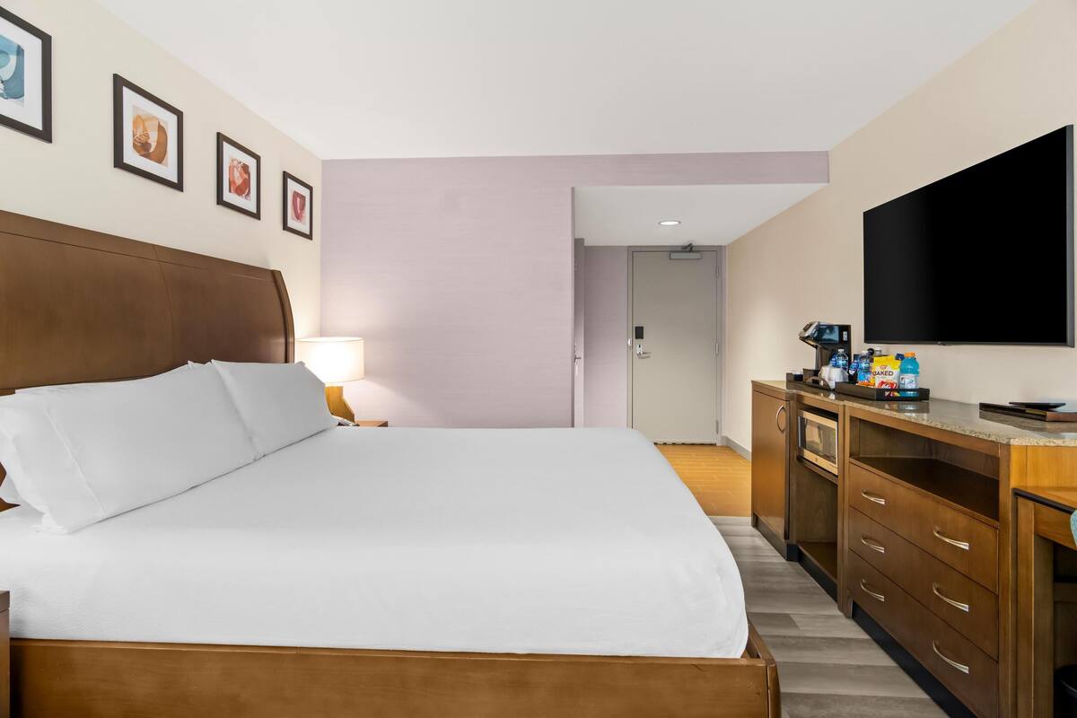 A hotel room with a large bed, wooden headboard, a flat-screen TV, a wooden dresser, and a door leading to another room.