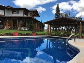 A swimming pool with a wooden deck and a covered area, surrounded by a well-maintained garden and a large house with multiple windows and balconies.