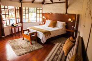 A bedroom with a wooden bed, a bedside table, a nightstand, a chair, a large window, and a view of the outdoors.