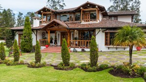 Une grande maison à deux étages, avec un toit en tuiles, des balcons en bois et un jardin agrémenté de chemins de pierre et de diverses plantes.