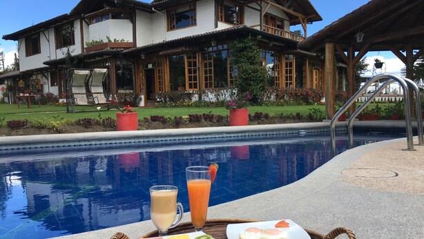Un plateau de petit-déjeuner avec des fruits, des œufs et des boissons, placé au bord de la piscine, et une grande maison en arrière-plan.