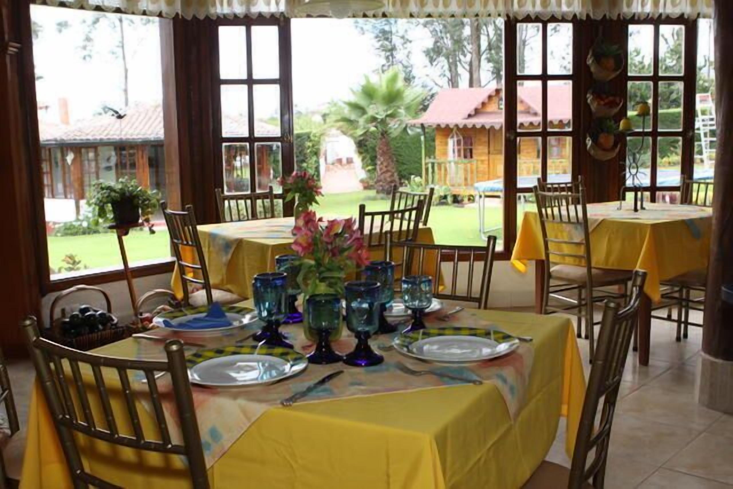 Un espace repas avec des tables dressées pour quatre personnes, chacune agrémentée d’une nappe jaune, de verres bleus et d’un vase avec des fleurs.