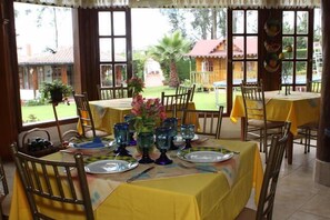 Un espace repas avec des tables dressées pour quatre personnes, chacune agrémentée d’une nappe jaune, de verres bleus et d’un vase avec des fleurs.