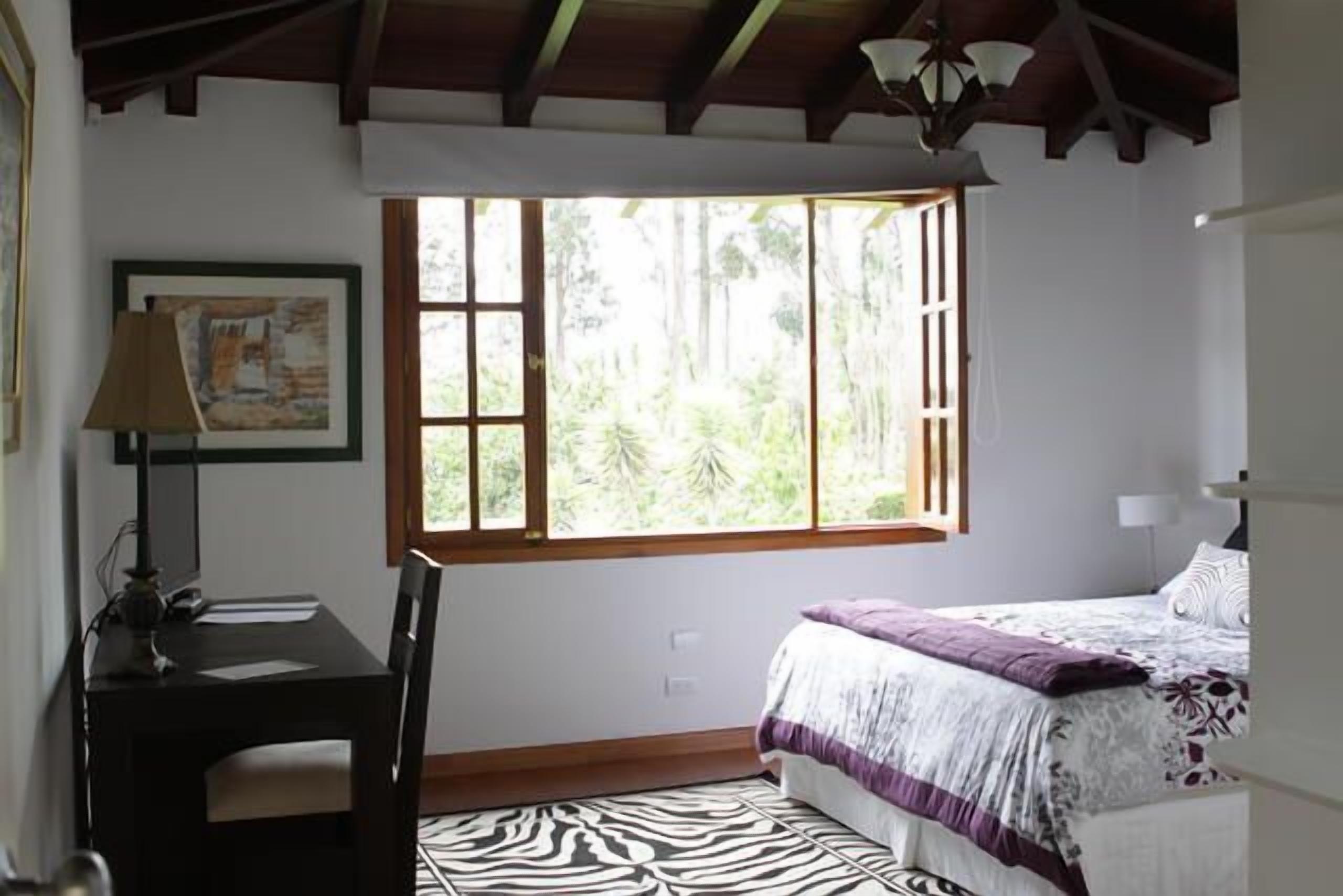 A bedroom with a bed, a desk, a chair, a window with a view of greenery, and a zebra-patterned rug.