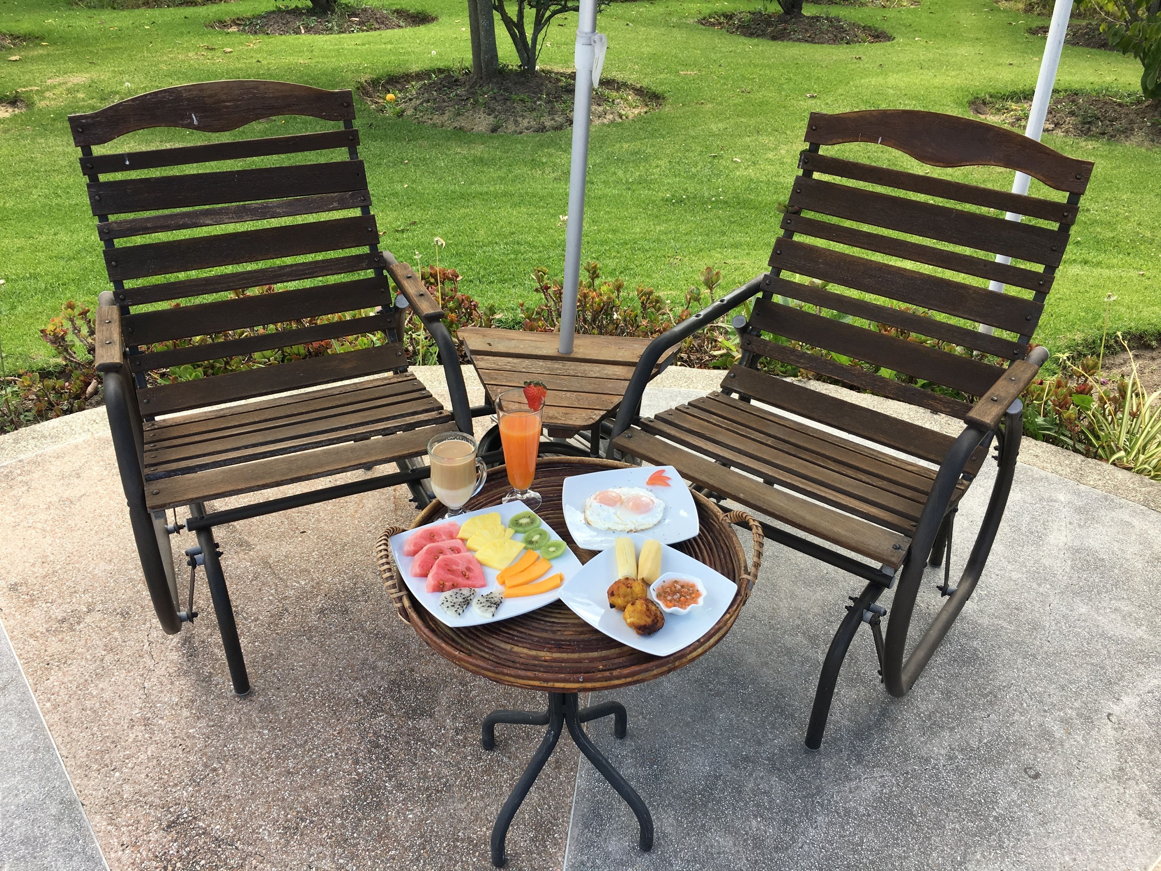 Two wooden chairs with metal frames and a round table with a wicker base. The table is set with plates of food, a glass of juice, and a cup of coffee.