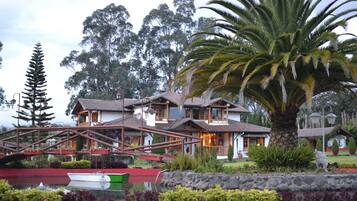 A traditional house with a wooden bridge, surrounded by lush greenery and a pond with a boat.