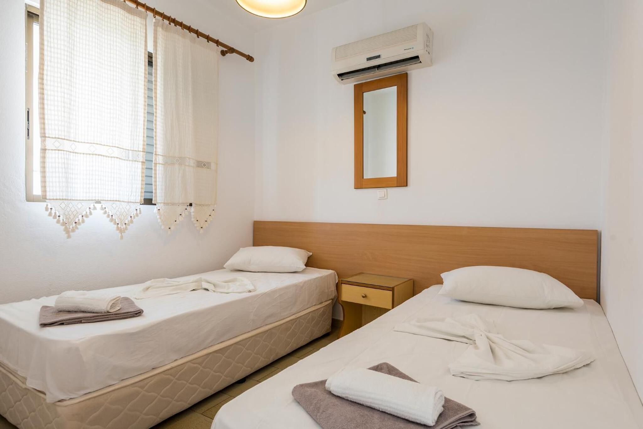 Habitación de hotel con dos camas individuales, cabecera de madera, mesita de noche, espejo y unidad de aire acondicionado.