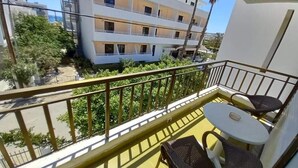 A balcony with a table and chairs, overlooking a building with multiple balconies and greenery. - Kardamena Holiday Apartments (Kos)