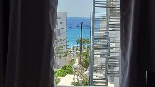 A view from a window with curtains, overlooking a coastal area with buildings and a clear sky. - Kardamena Holiday Apartments (Kos)