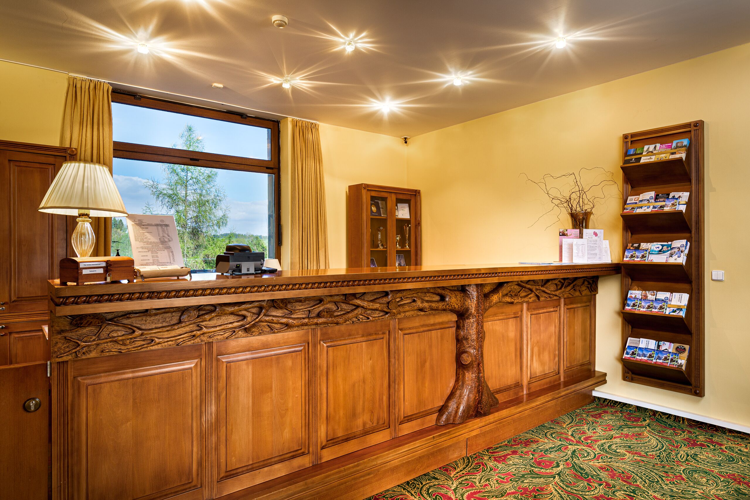 A wooden reception desk with a lamp, a bulletin board, and a bookshelf with brochures.