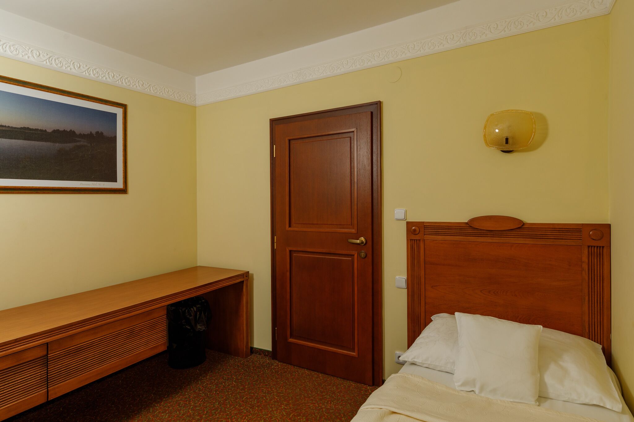 Habitación de hotel con una cama de madera, un banco, una puerta y un aplique en la pared.