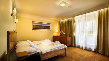 A hotel room with a bed, a desk, a lamp, a vase with flowers, a painting on the wall, and a window with curtains.