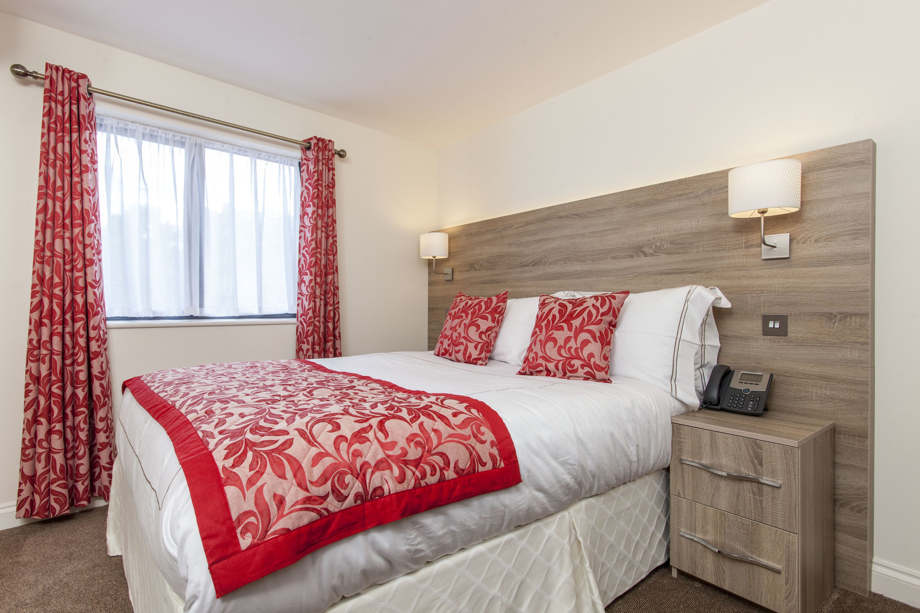 A neatly made bed with a red and white patterned bedspread, a wooden headboard, bedside lamps, and a bedside phone.