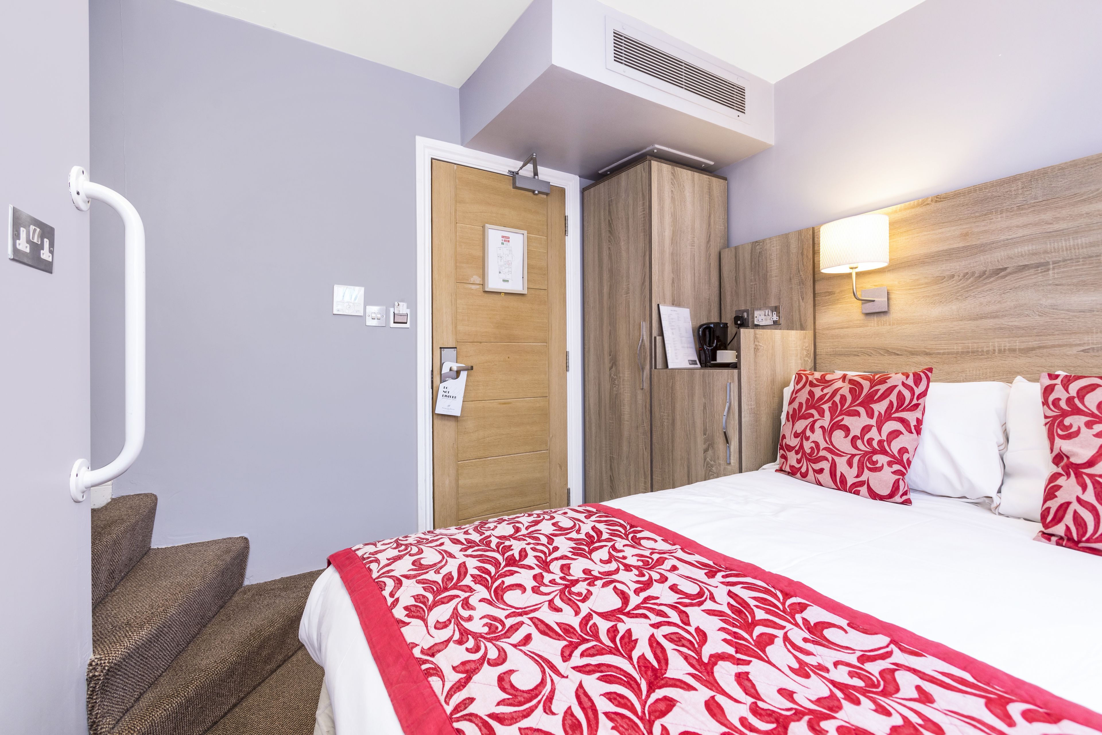 A hotel room with a bed, a wooden wardrobe, a white wall, a red and white patterned bedspread, a white bedside table, and a white lamp.