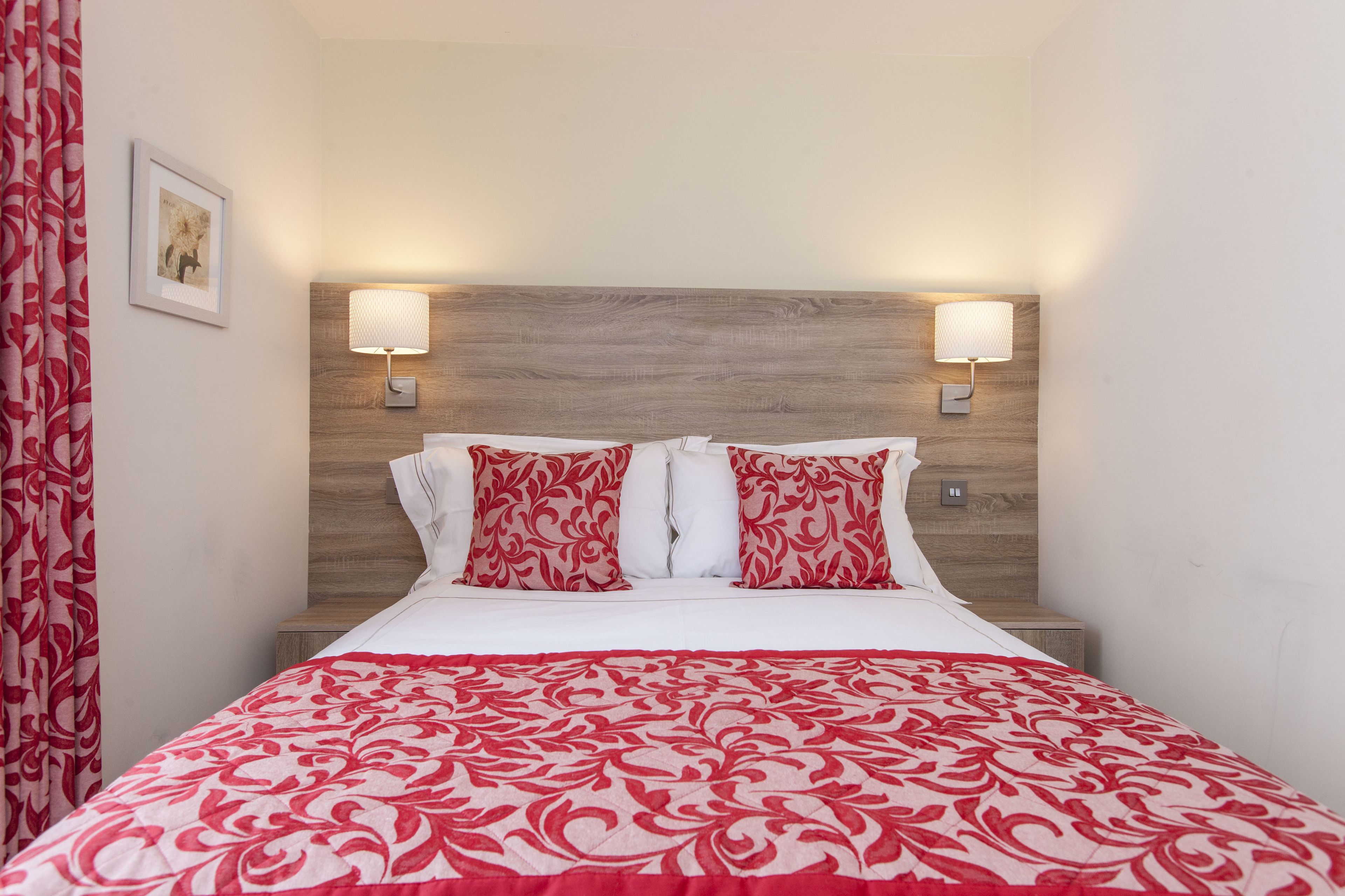 A hotel room with a wooden headboard, two bedside lamps, a bed with a red and white patterned bedspread, and a framed picture on the wall.