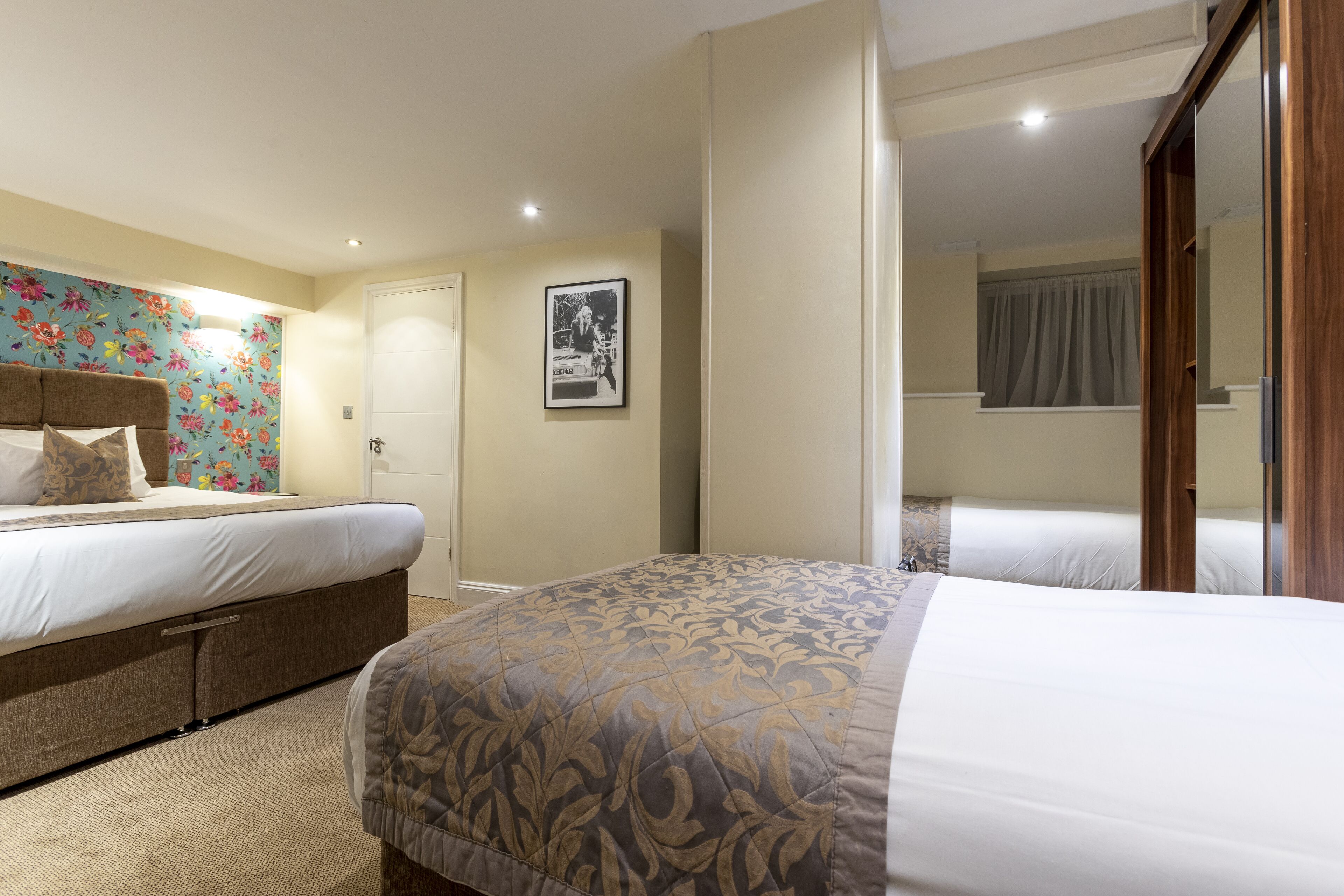 A hotel room with two beds, a floral wallpaper on one wall, a framed picture, and a wooden wardrobe.