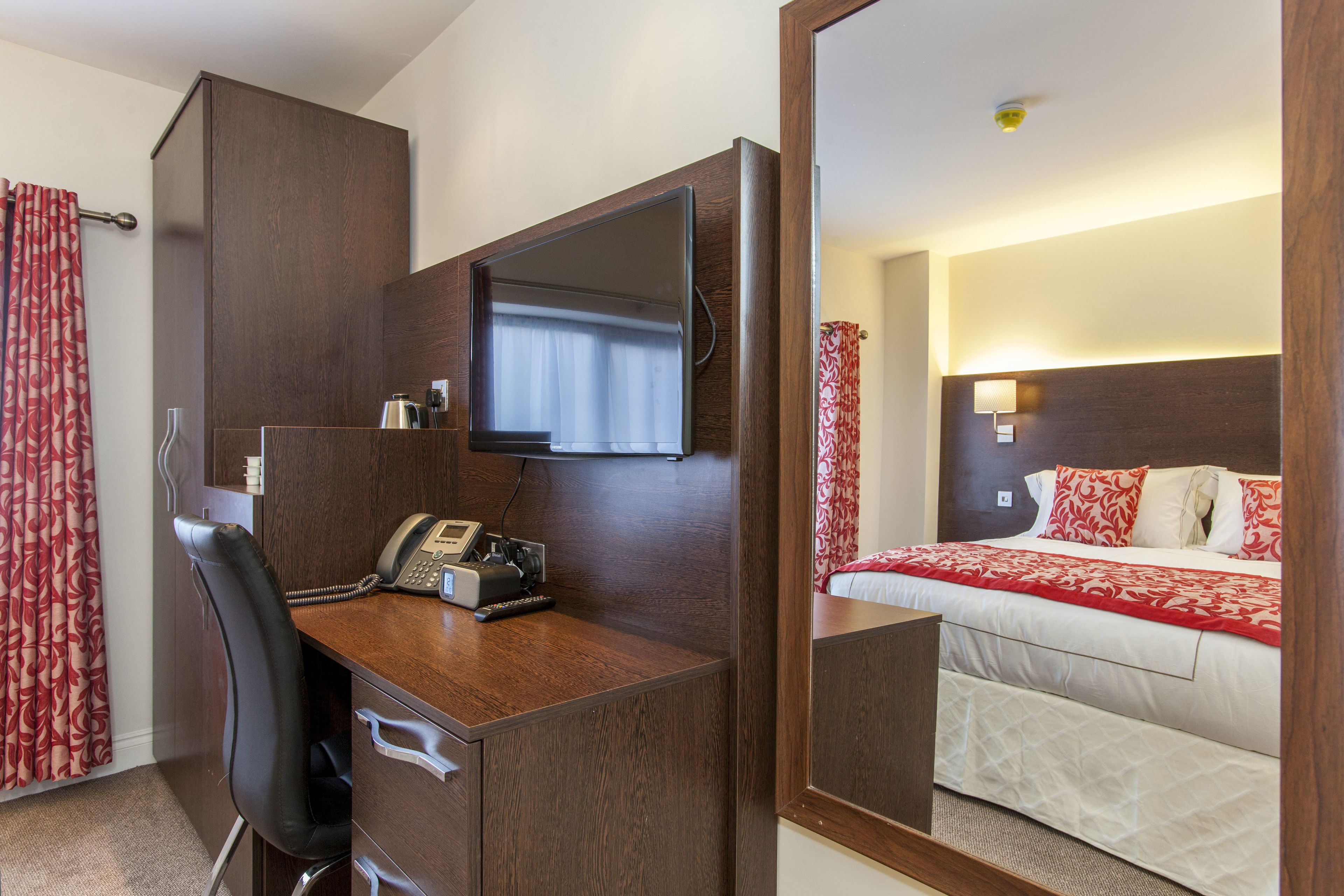 A hotel room with a wooden desk, a chair, a bed with red and white bedding, and a large mirror.