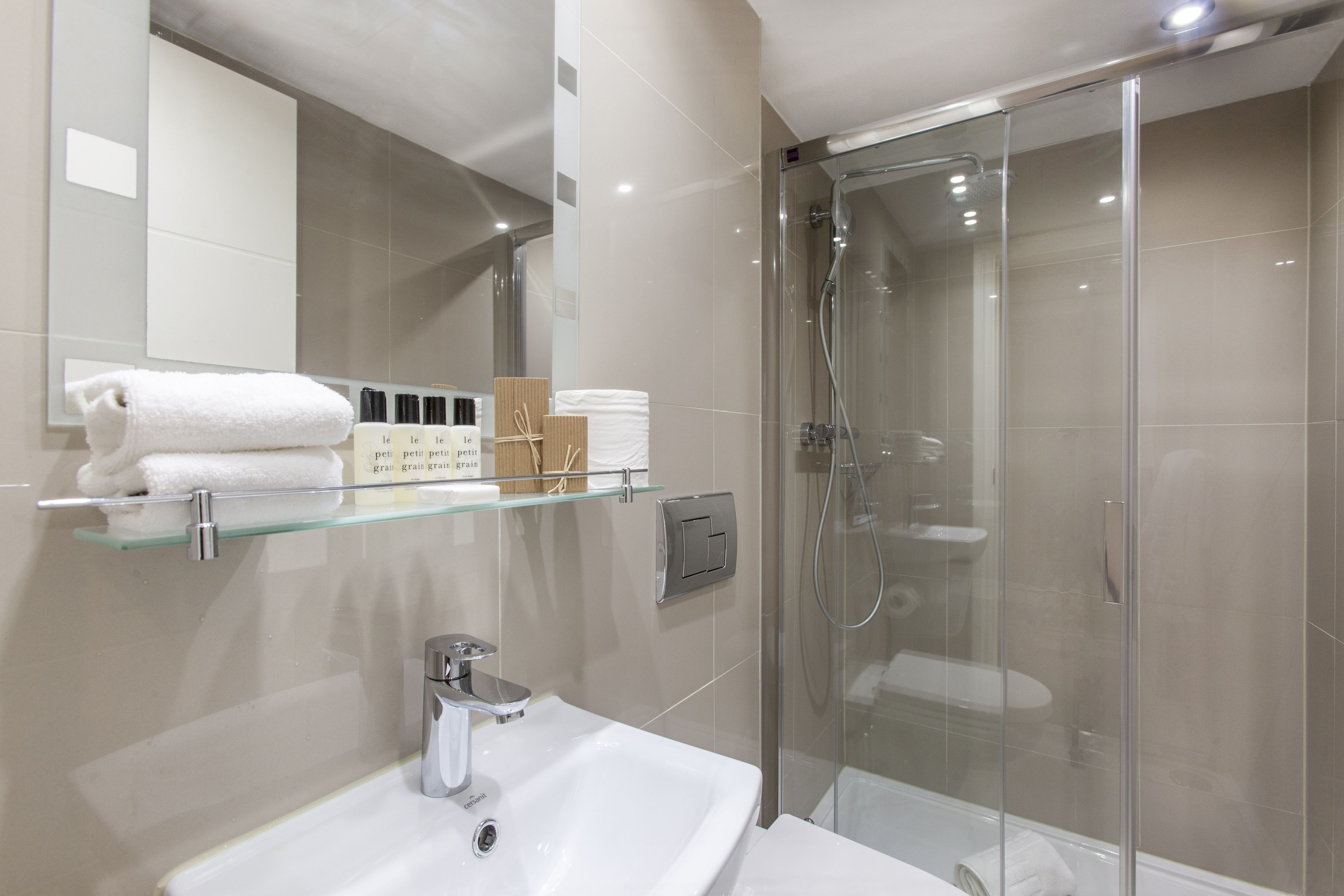 Et moderne badeværelse med en glasvæg omkring brusebadet, en hvid vask og en glashylde med toiletartikler.