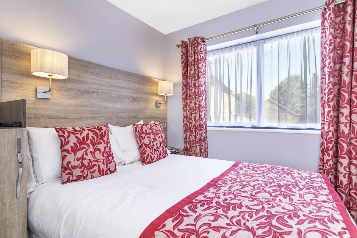 A hotel room with a bed, two wall-mounted lamps, a window with curtains, and a wooden headboard.
