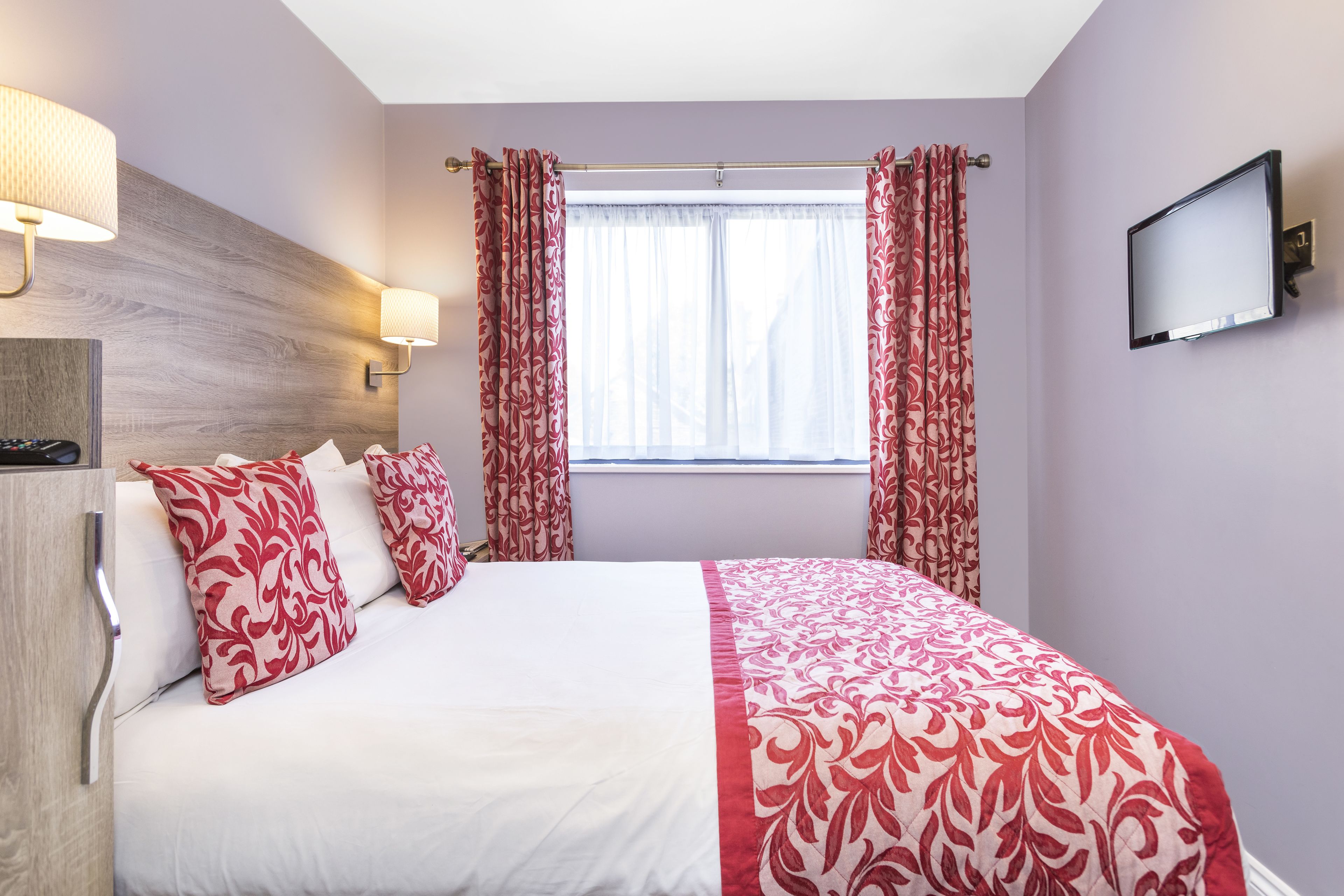 A hotel room with a bed, two pillows, a nightstand, a TV, and a window with curtains.