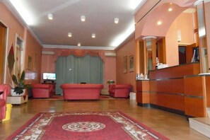A hotel lobby with a reception desk, seating area with sofas, a television, and a patterned carpet. - Hotel Alpen (Rimini)