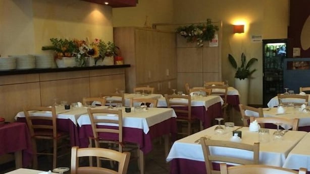 Un restaurant doté de nombreuses tables et chaises, chacune recouverte d’une nappe blanche et agrémentée d’un chemin de table bordeaux. Des plantes en pot décorent les murs, et une étagère est garnie d’assiettes empilées.
