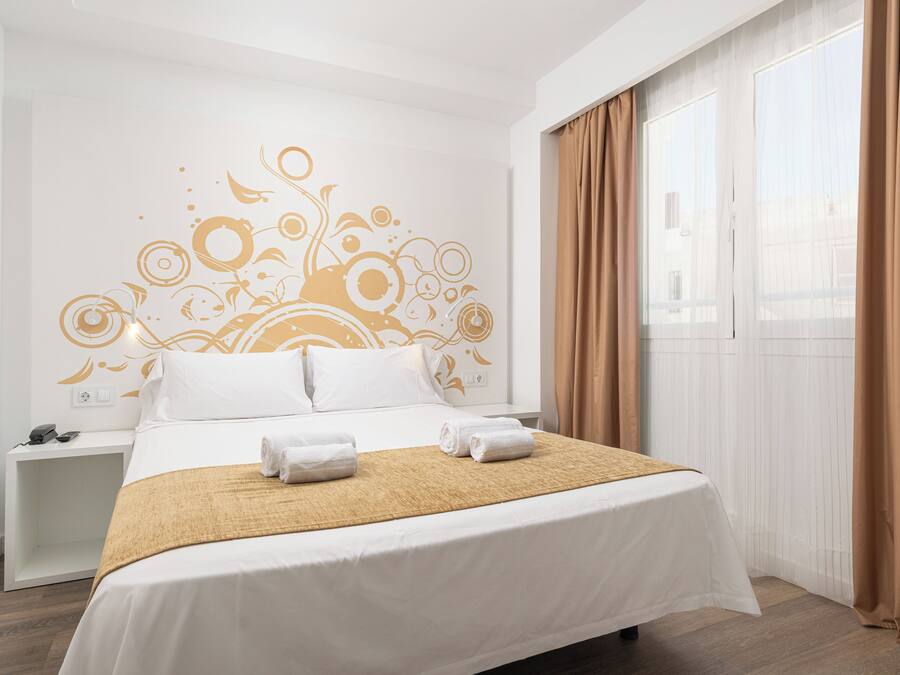 Una habitación de hotel con cama, mesita de noche, decoración en la pared y una ventana con cortinas.