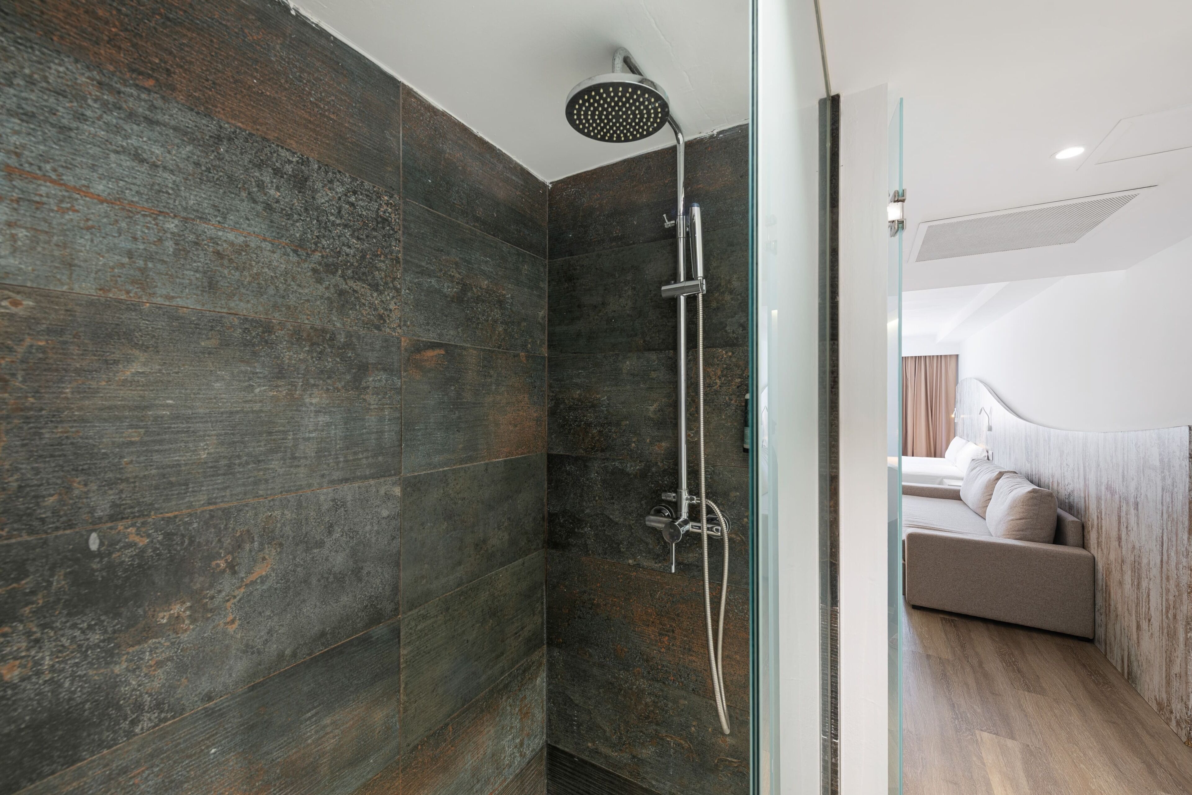 Un baño moderno con ducha, puerta de vidrio y vista hacia un dormitorio con cama y mesita de noche.