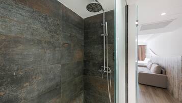 Un baño moderno con ducha, puerta de vidrio y vista hacia un dormitorio con cama y mesita de noche.