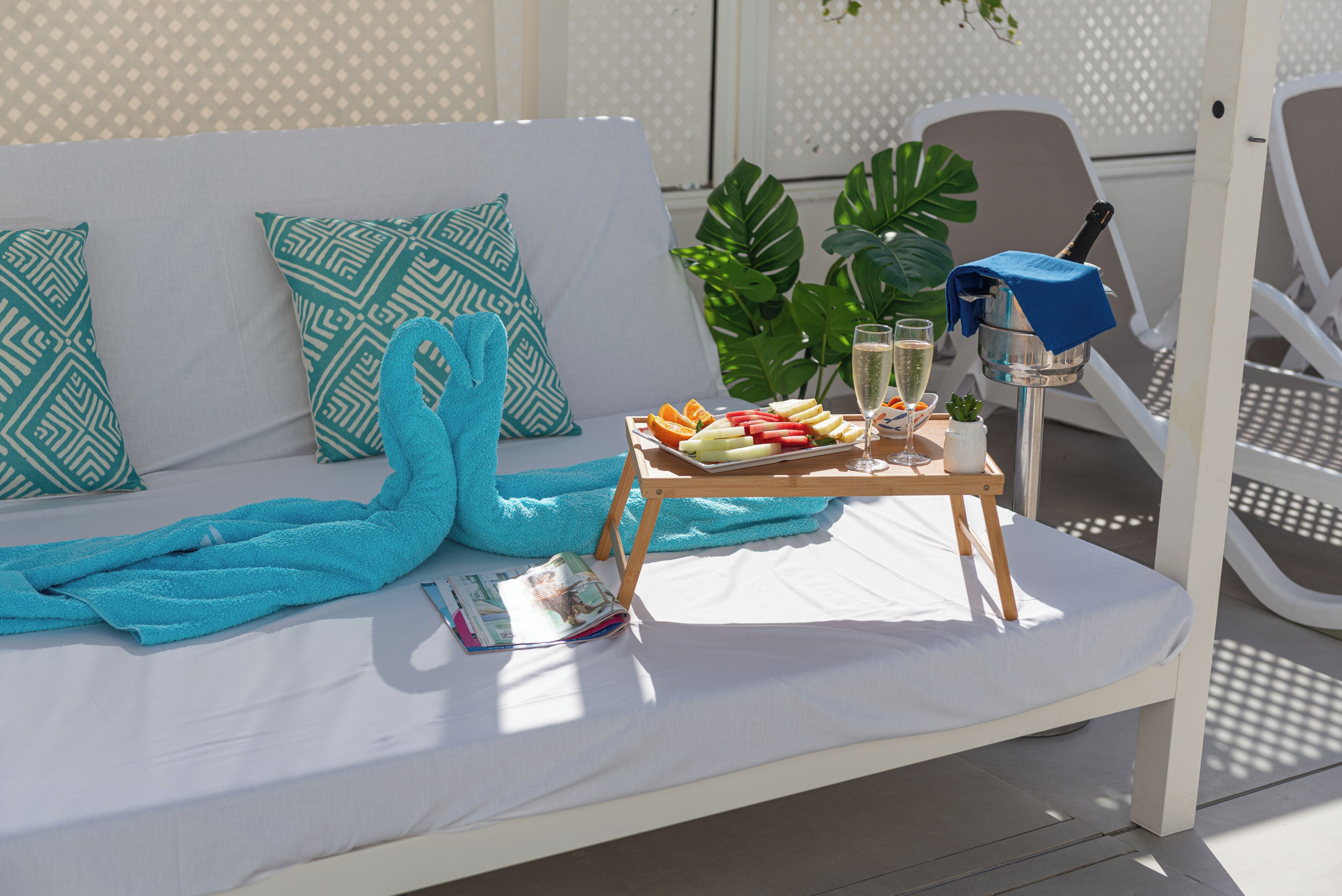 A sunlit outdoor seating area with a white sofa, blue towels, a tray with fruits and a drink, and a blue umbrella.