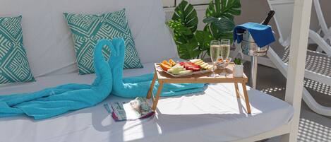A sunlit outdoor seating area with a white sofa, blue towels, a tray with fruits and a drink, and a blue umbrella.