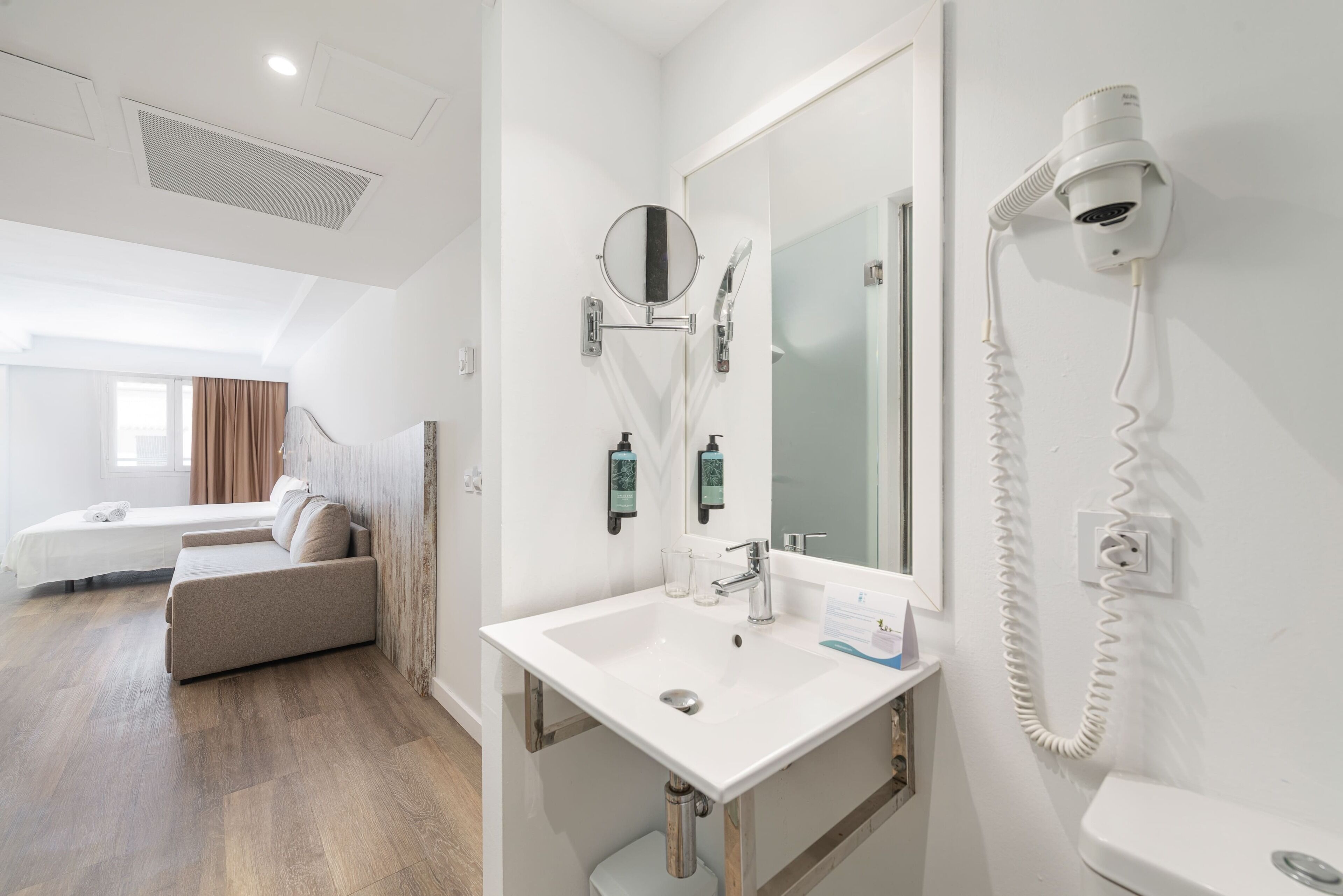 Habitación de hotel con cama, sofá, baño con lavamanos, espejo y secador de pelo.