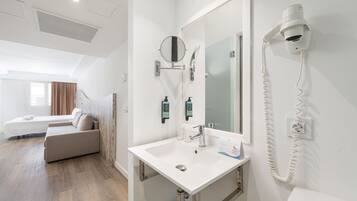 Habitación de hotel con cama, sofá, baño con lavamanos, espejo y secador de pelo.