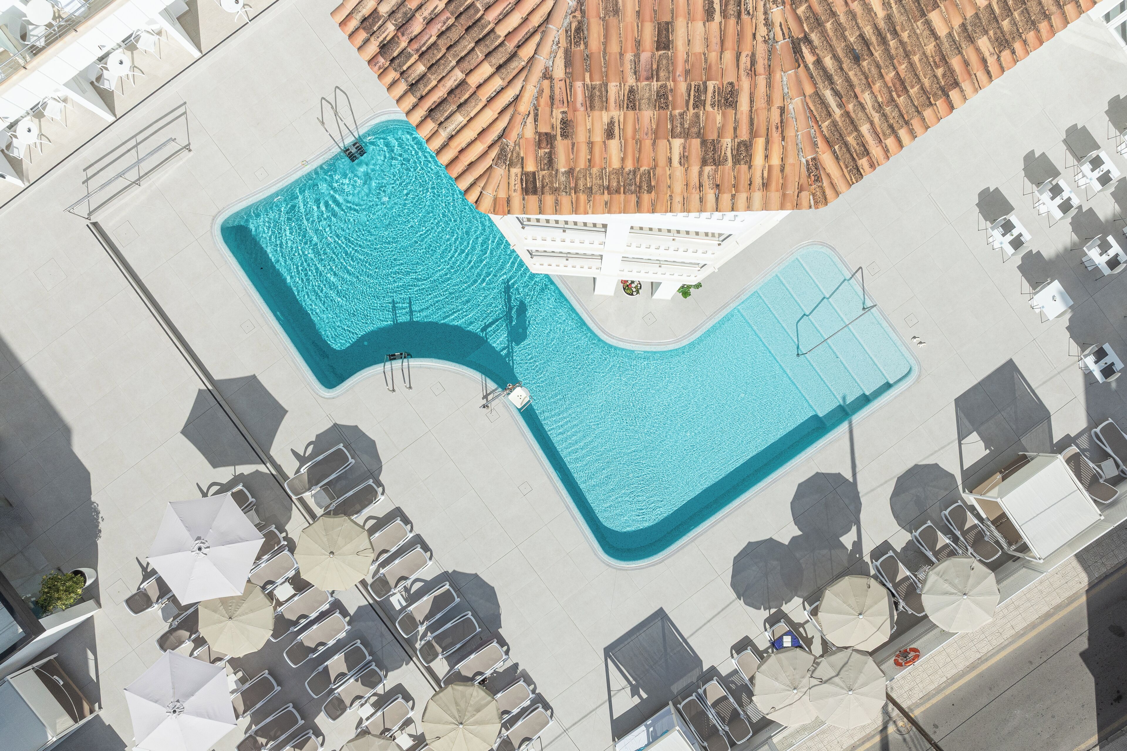 An aerial view of a hotel with a large swimming pool, multiple lounge areas with chairs and umbrellas, and a tiled roof.