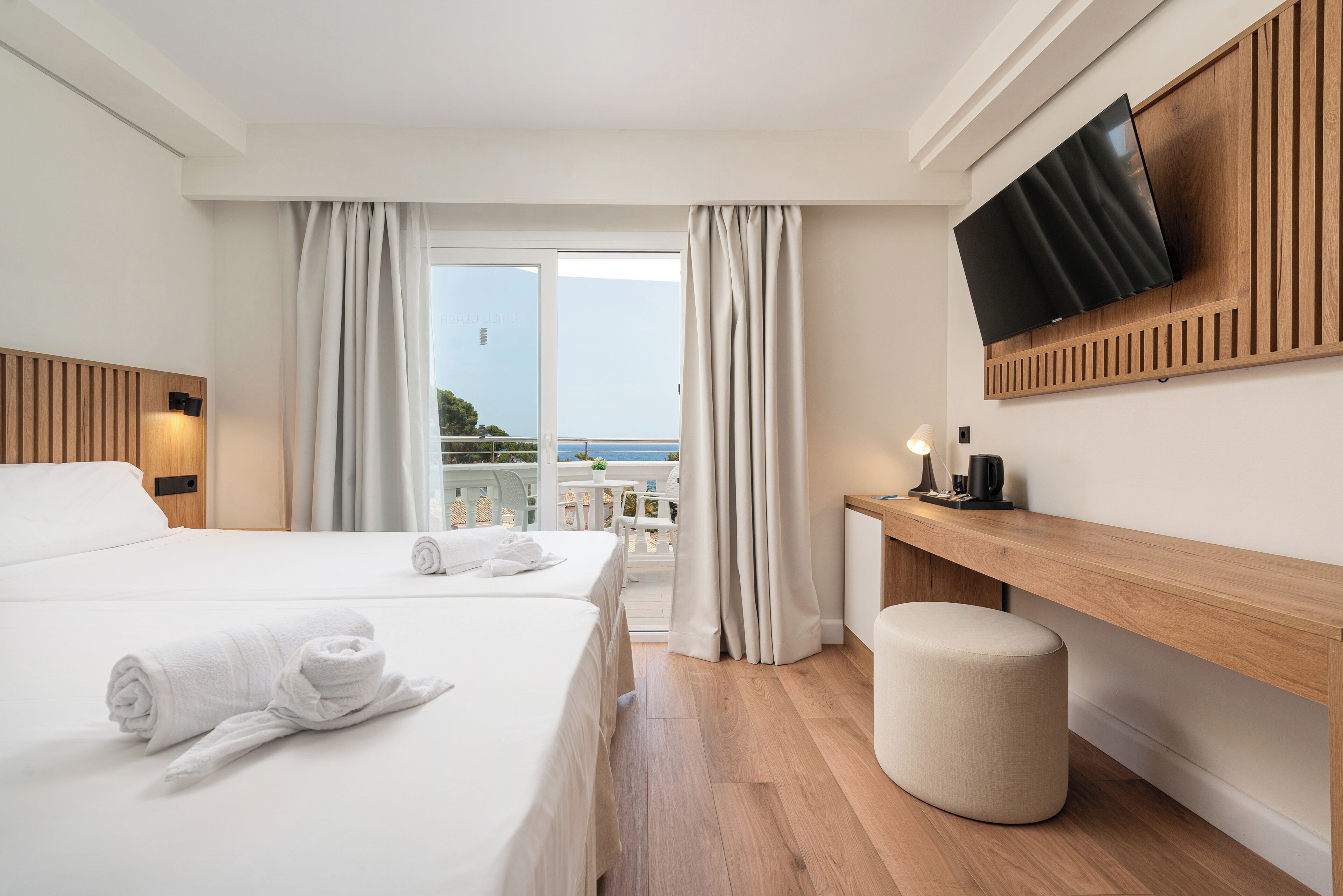A hotel room with a bed, a desk, a TV, and a view of the sea.