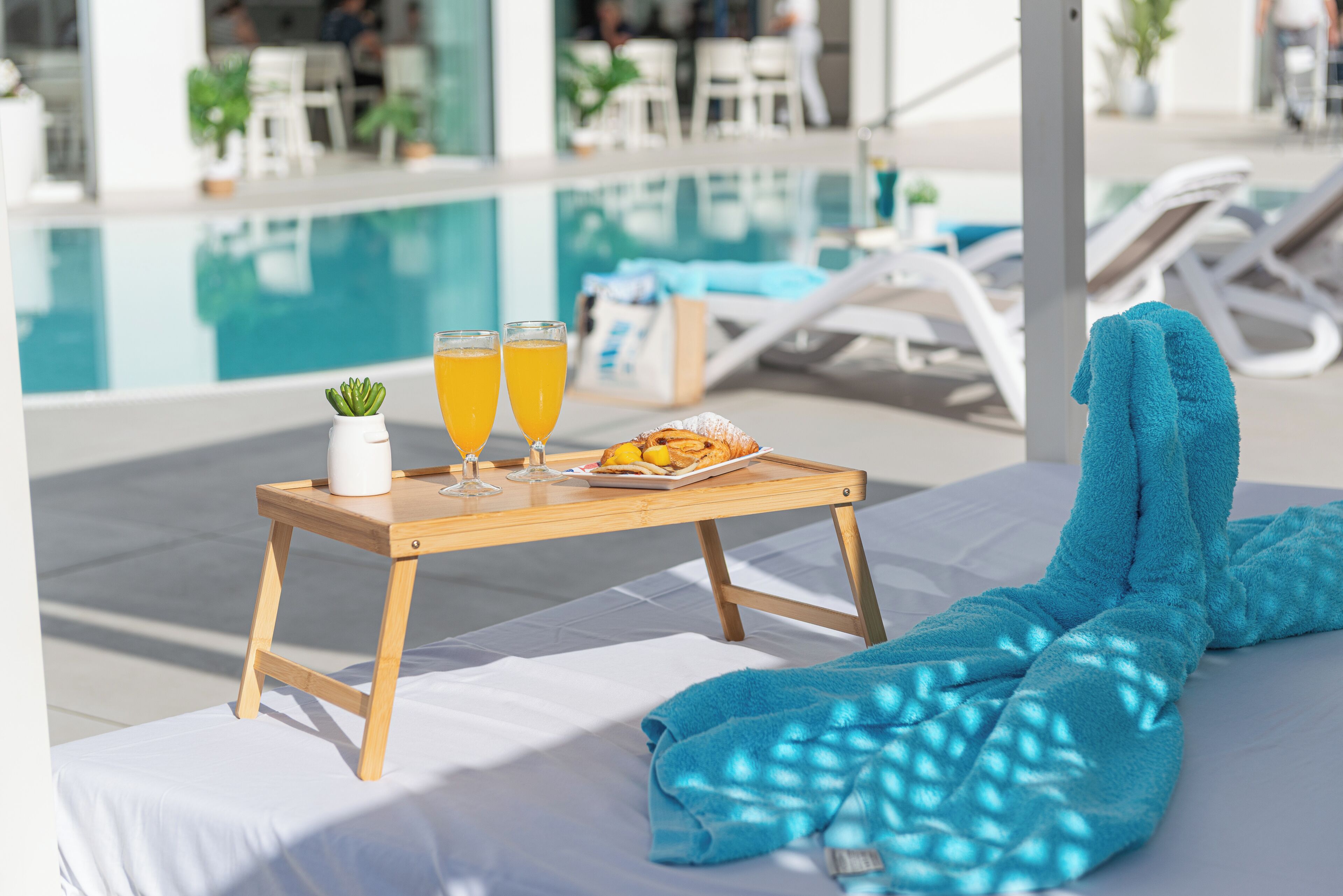 Cenário junto à piscina, com uma bandeja de madeira que contém um prato de comida, dois copos de suco de laranja e uma pequena planta em vaso.