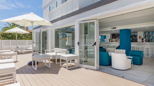 Una terraza al aire libre con mobiliario blanco y azul, una zona de bar y una entrada con puerta de cristal.