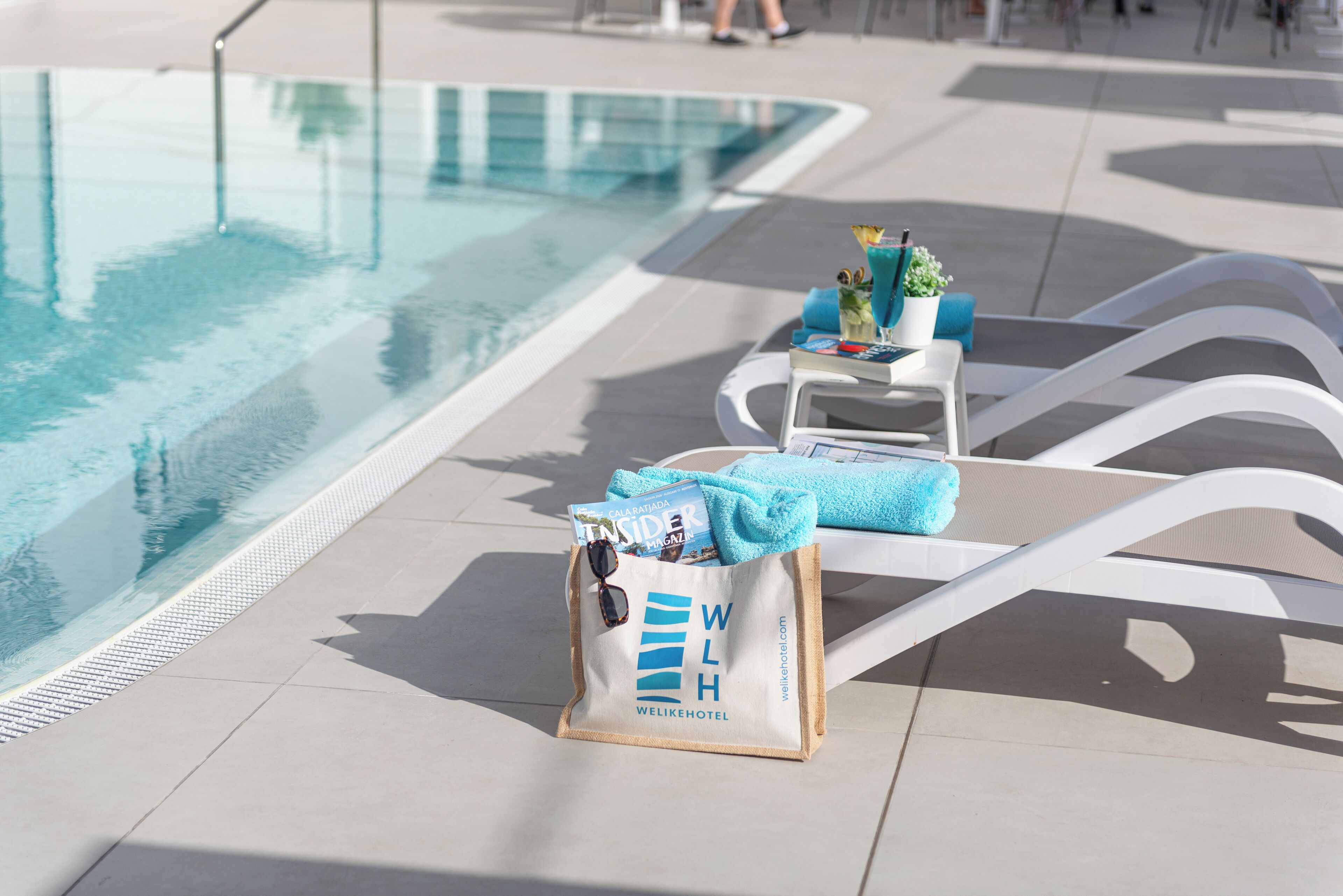 A poolside area with lounge chairs, a bag with the hotel logo, and a clear pool.