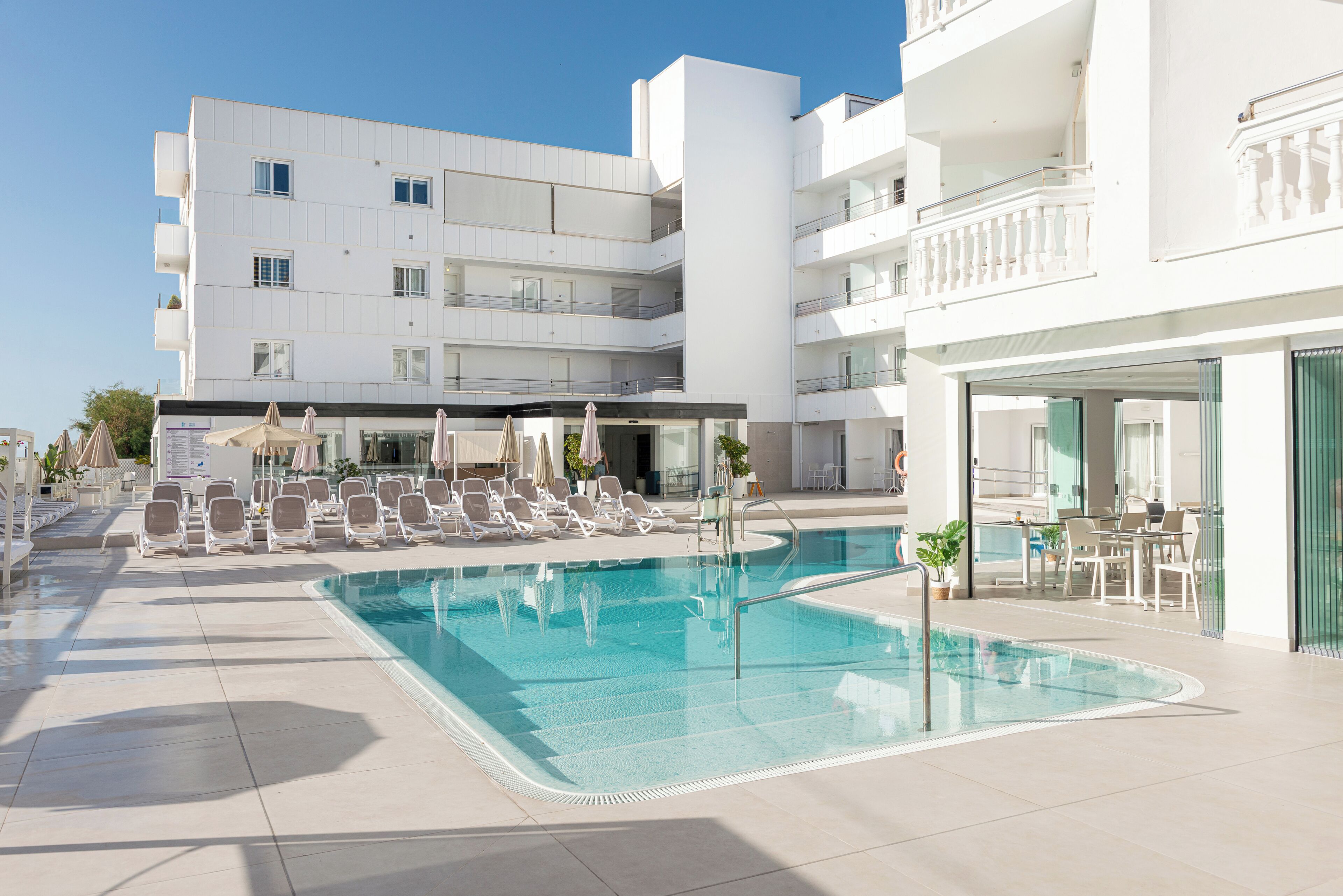 A pool area with lounge chairs and umbrellas, a clear sky, and a white building with multiple balconies.