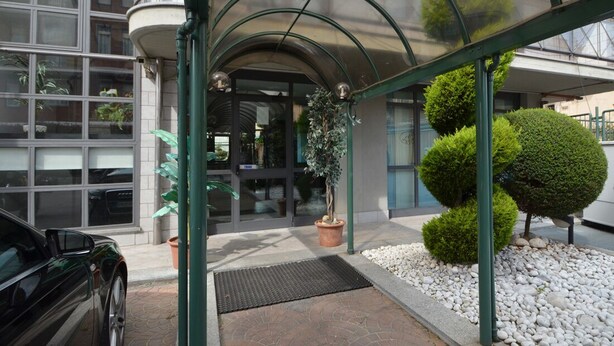 A building entrance with a covered walkway, potted plants, and a car parked outside. - Hotel delle Alpi (Turin)