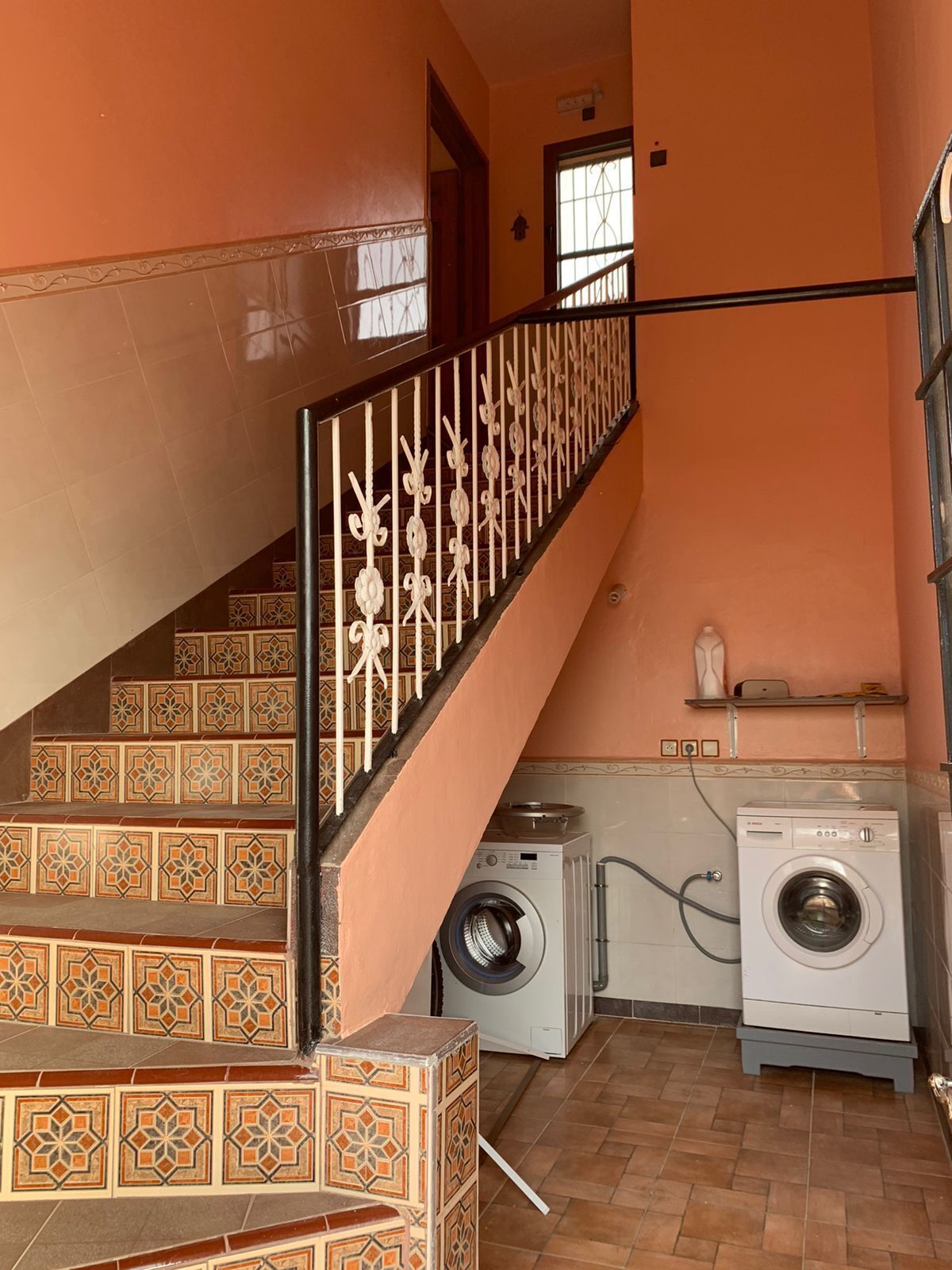 Un área de lavandería con dos lavadoras, una escalera con baldosas estampadas y una ventana con celosía decorativa.