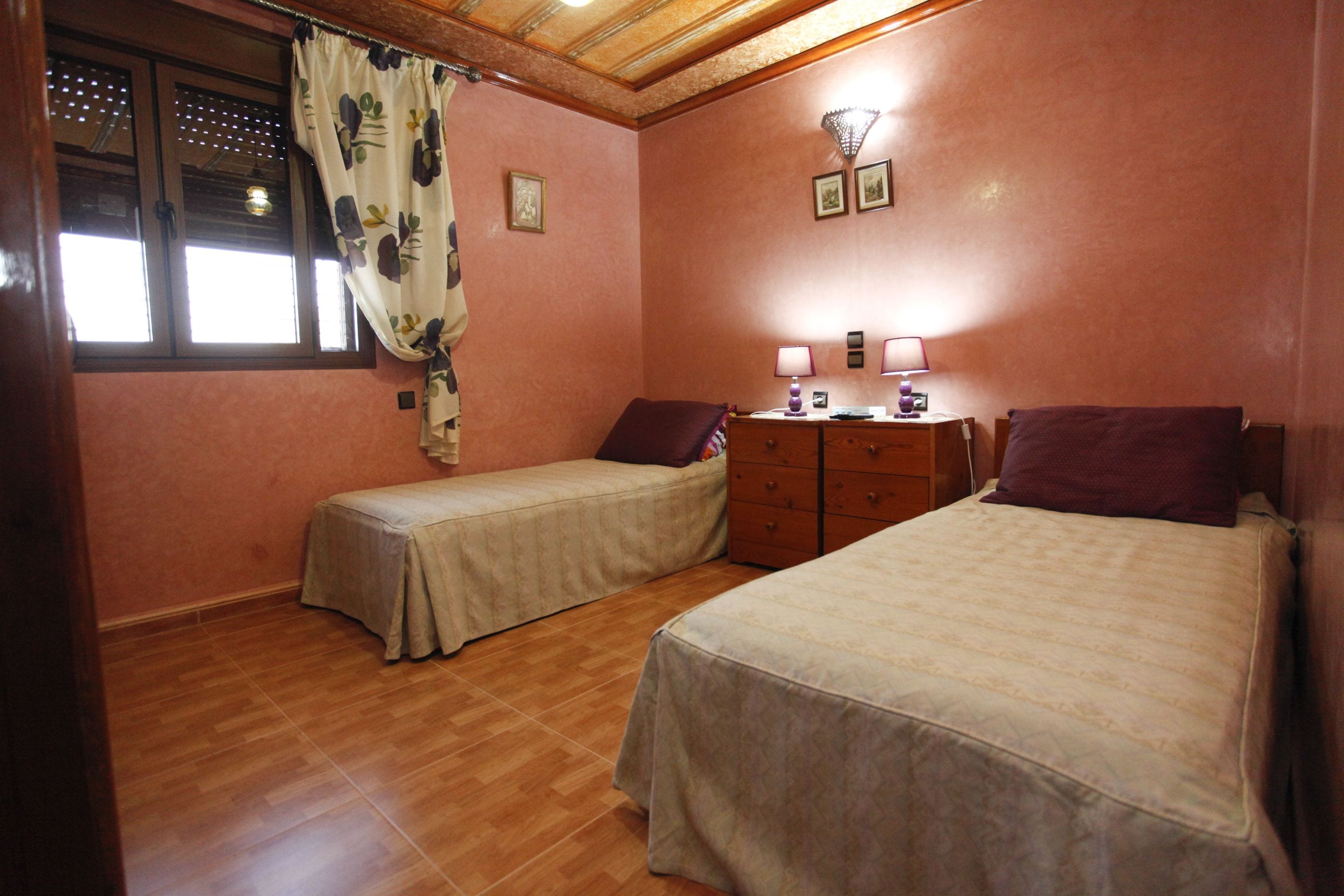 A bedroom with two single beds, a wooden dresser, a nightstand, and a window with curtains.