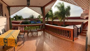 A covered outdoor area with a table and chairs, a wooden railing, and a view of palm trees and buildings.