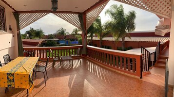 Un espacio al aire libre cubierto con una mesa, sillas, baranda de madera y vistas a palmeras y edificios.
