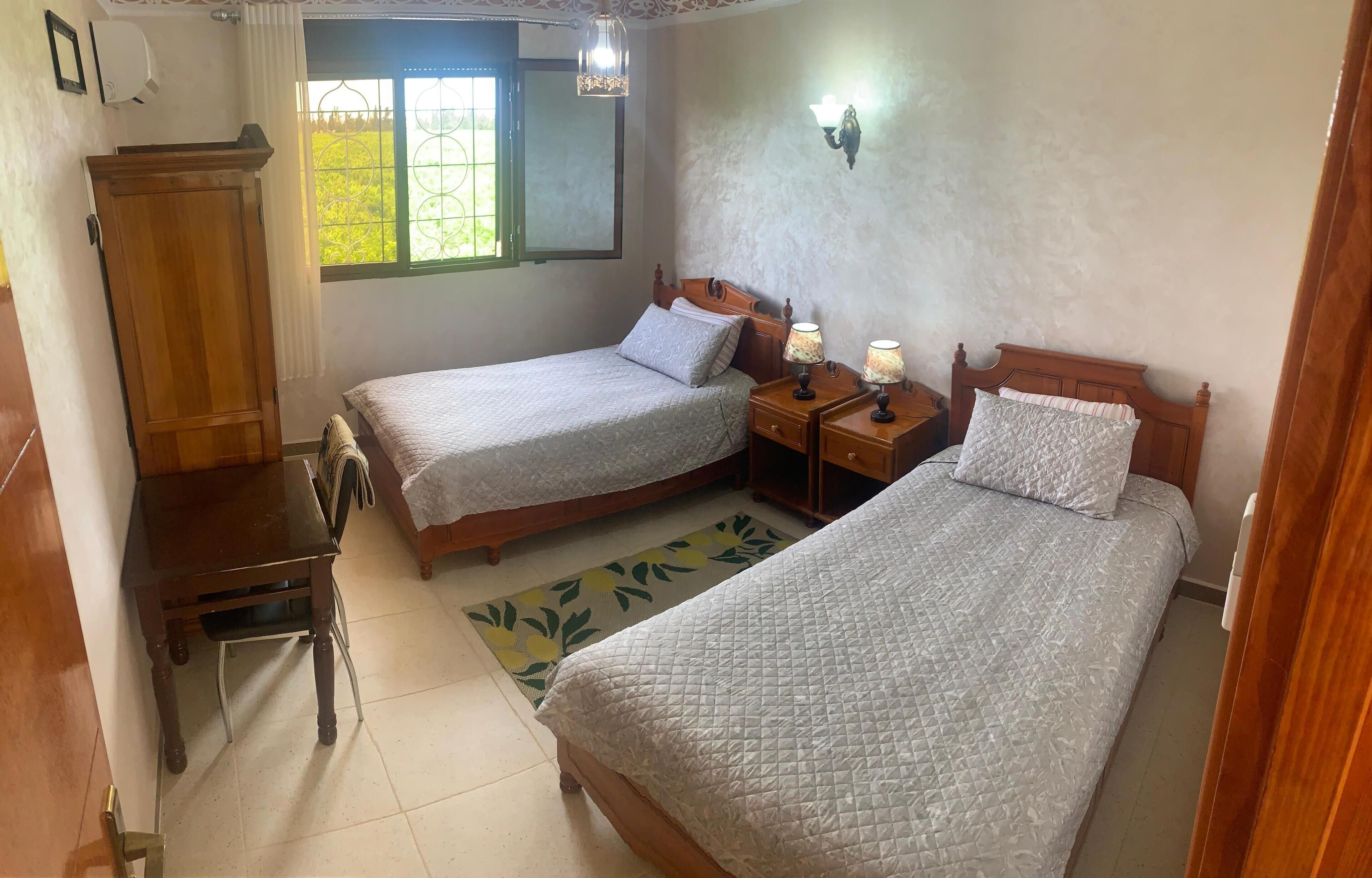 A bedroom with two single beds, a wooden wardrobe, a small table, and a nightstand with lamps.
