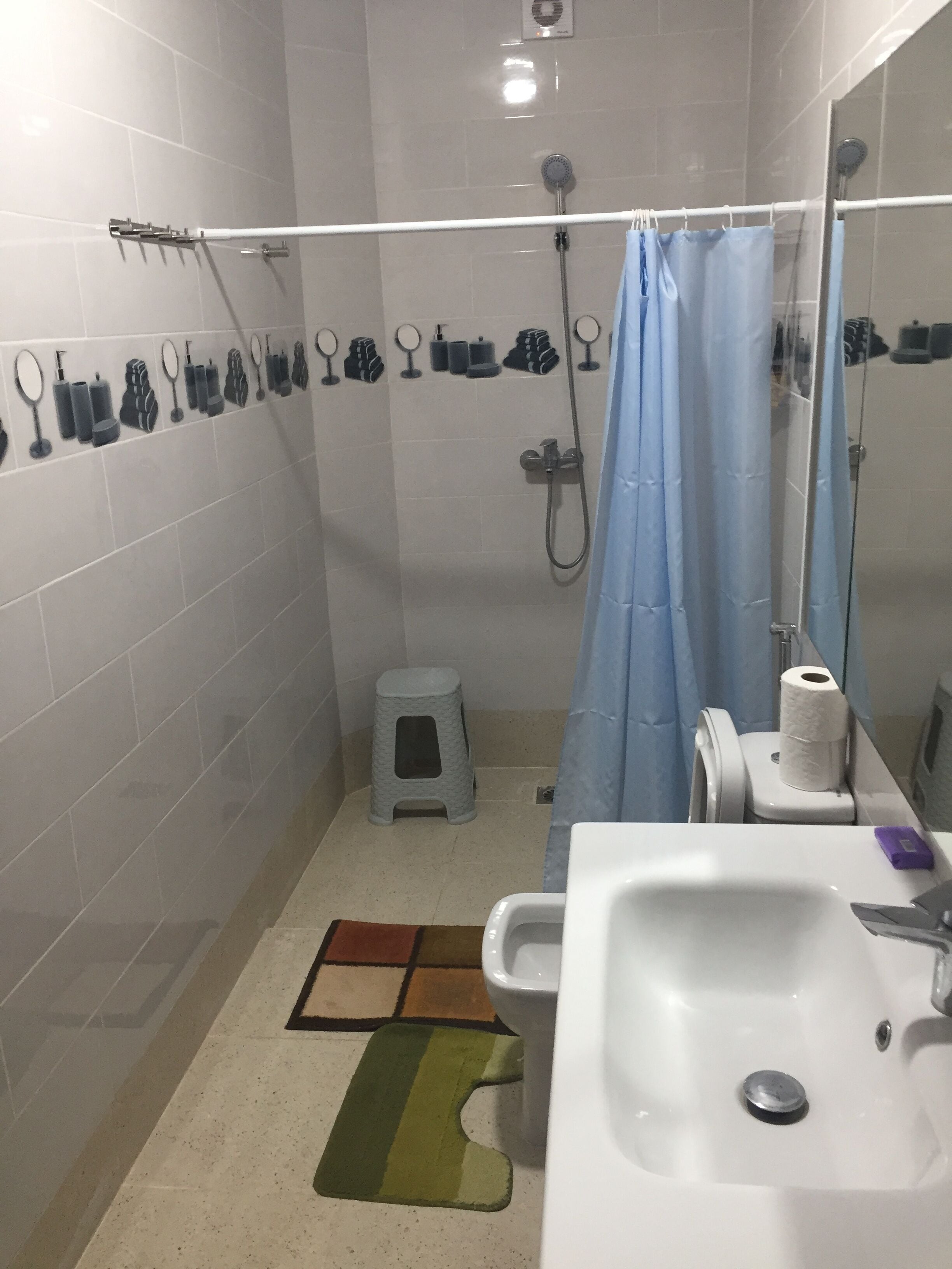 Un baño con ducha, inodoro y lavamanos. Hay un taburete pequeño y un perchero con distintos artículos de aseo.