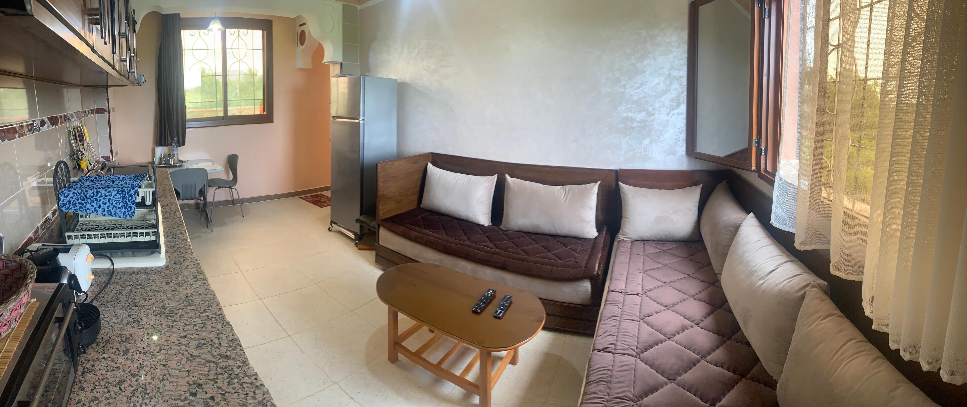 Una sala de estar con sofá, una pequeña mesa de madera, una zona de cocina con refrigerador y una ventana con cortinas.