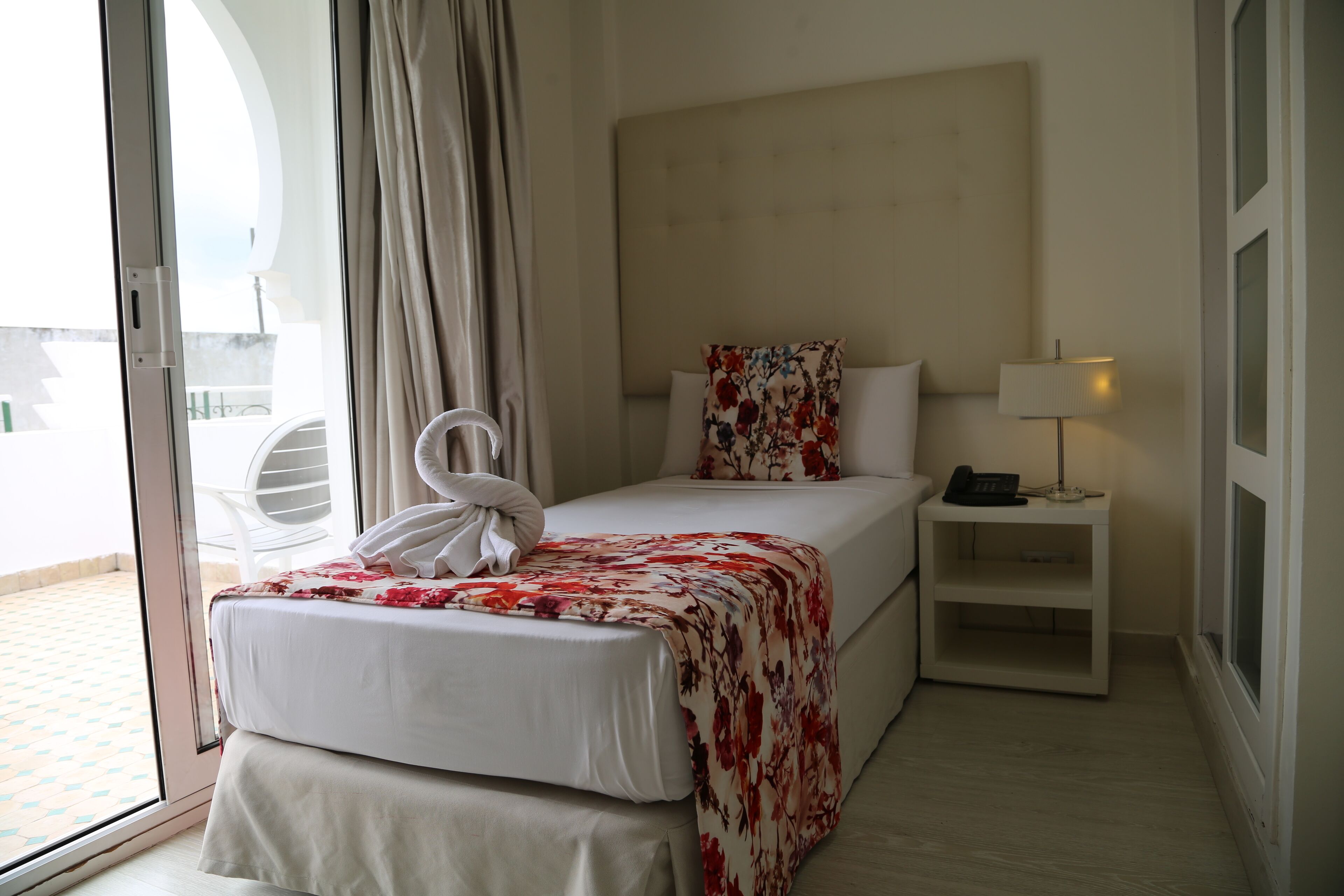 A hotel room with a bed, a bedside table with a lamp, and a view through a glass door.